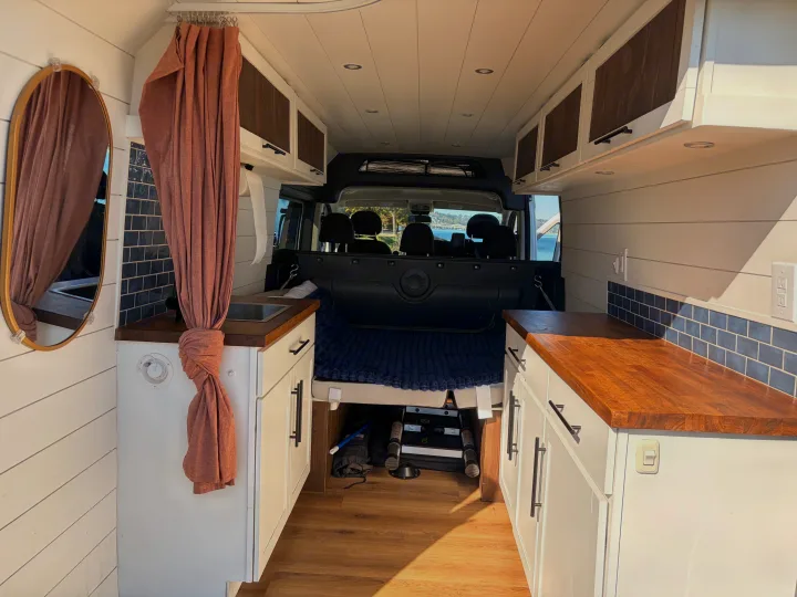 RV rental's cozy interior: bed, compact kitchen with wood counters, and white cabinets for comfortable van life adventures.. Dodge RAM Promaster 3500 2020
