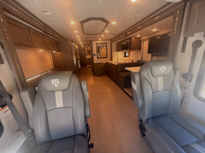 Modern RV rental interior with grey captain's chairs, wood floor, full kitchen, and a dining area visible towards the rear.. Thor Riviera 2024