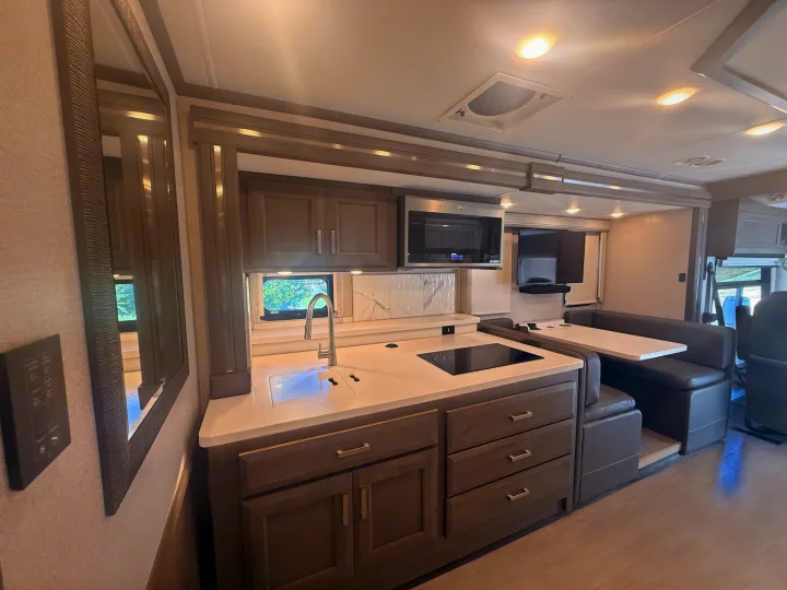 Modern rv rental interior featuring a compact kitchen with sink and stovetop, built-in dinette, and wall-mounted TV.. Thor Riviera 2024