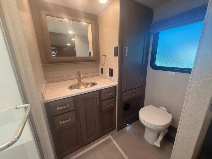 This compact RV rental bathroom features a mirrored vanity, sink, toilet, and shower handle, offering complete facilities for travelers.. Thor Riviera 2024