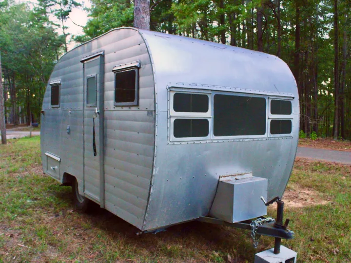 A classic silver travel trailer, available as an rv rental, is parked outdoors in a wooded area, offering a rustic escape.. Shasta 1500 1954