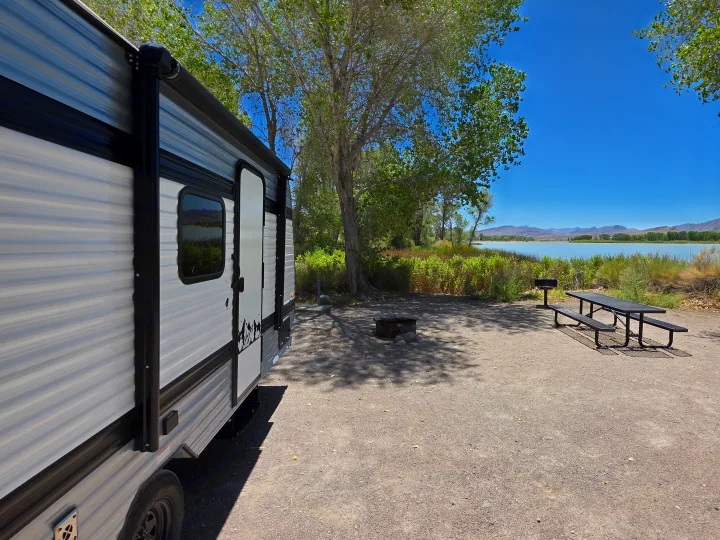 RV rental at a scenic lakeside campsite with a picnic table and fire pit, surrounded by trees under a clear blue sky.. Keystone Coleman 17B 2025