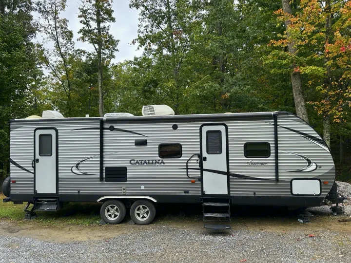Gray Coachmen Catalina RV rental parked amidst trees. Features two doors, extended steps, and stabilizers for camping.. Coachmen Catalina 2017