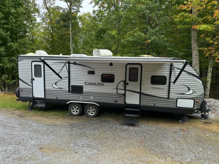 This Catalina travel trailer rv rental sits on a gravel campsite amidst lush trees, perfect for a peaceful outdoor escape.. Coachmen Catalina 2017