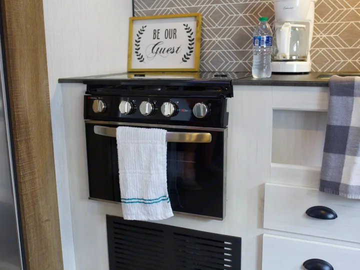 "Be Our Guest" in this RV rental kitchen, featuring a compact gas stove, oven, coffee maker, and checkered decor.. Heartland Trail Runner 2022