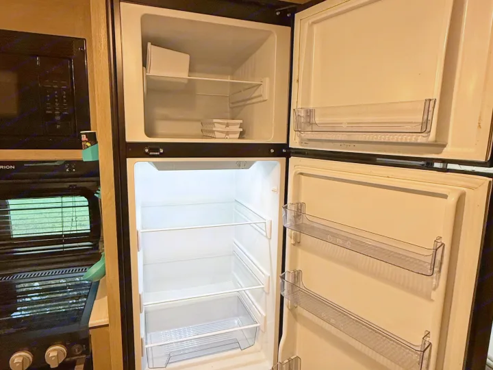An RV rental kitchen with an open, empty refrigerator and freezer showing shelves, door bins, a microwave, and stovetop.. Forest River Wildwood X-Lite Platinum 2024