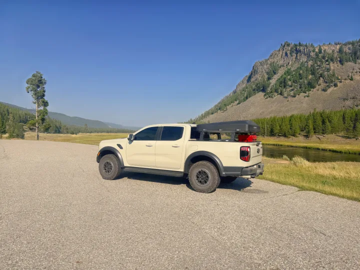 Adventure-ready pickup truck with rooftop tent, a rugged RV rental alternative, parked by a scenic river in a mountain landscape.. Ford Ranger 2025