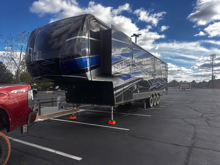 A large, black and blue "NITRO" fifth-wheel RV (rv rental) parked in a lot, with a red pickup truck & Oklahoma Sooners trailer.. Forest River XLR Nitro Toy Hauler 2023