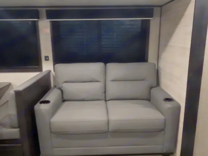 RV rental interior featuring a gray loveseat with cup holders, dark window blinds, and an exit sign for safe travels.. Jayco Jay Flight 2023