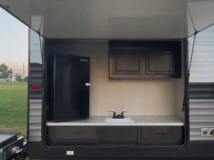 This RV rental's outdoor kitchen features a black mini-fridge, sink, countertop, and cabinets, providing convenience for outdoor meals.. Jayco Jay Flight 2023
