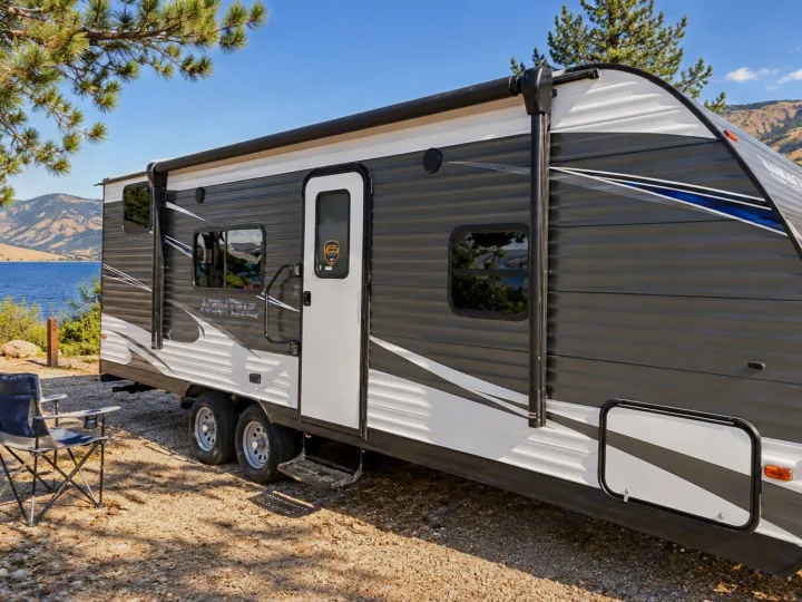 This RV rental, a stylish travel trailer, is set up at a scenic lakeside campsite under a clear sky. Perfect for outdoor adventures.. Dutchmen Aspen Trail 2020