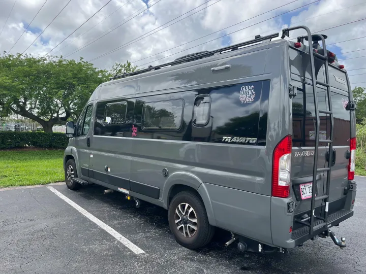 This gray Winnebago Travato RV rental, with a roof rack and rear ladder, is parked in a lot on a cloudy day.. Winnebago Travato 2024