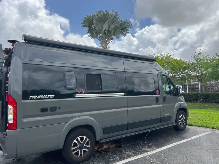 Grey Winnebago Travato rv rental parked outdoors, a dog rests under it. Blue sky & palm tree overhead.. Winnebago Travato 2024
