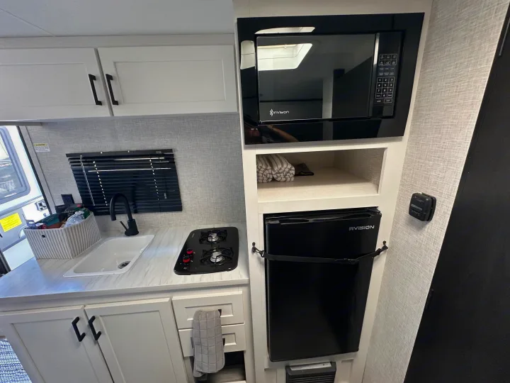 An RV rental's compact kitchen features white cabinets, microwave, sink, two-burner stove, and small fridge.. Jayco Jay Flight 2025