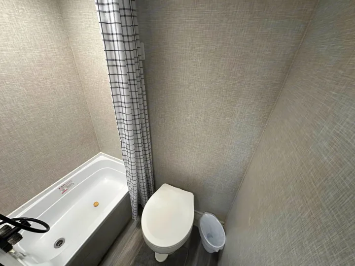RV rental bathroom featuring a compact tub/shower combo with plaid curtain, toilet, and textured light gray walls.. Jayco Jay Flight 2025