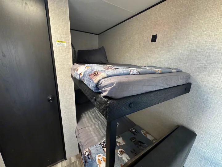 Inside an RV rental: a compact bunk bed setup with cozy dog blankets, grey bedding, and a USB charging port.. Jayco Jay Flight 2025