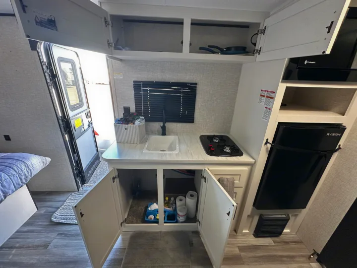 Inside an RV rental: compact kitchenette with sink, two-burner stove, mini-fridge, and open cabinets showing storage. Door and bed visible.. Jayco Jay Flight 2025
