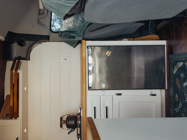 A top-down view of an RV rental interior: a compact kitchen area with a stainless Frigidaire mini-fridge, white cabinetry, and a kettle.. Ford Transit 2015