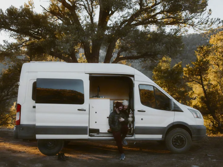 A happy couple and their dog relax in a white camper van with the door open, surrounded by trees. Ideal for an RV rental adventure!. Ford Transit 2015