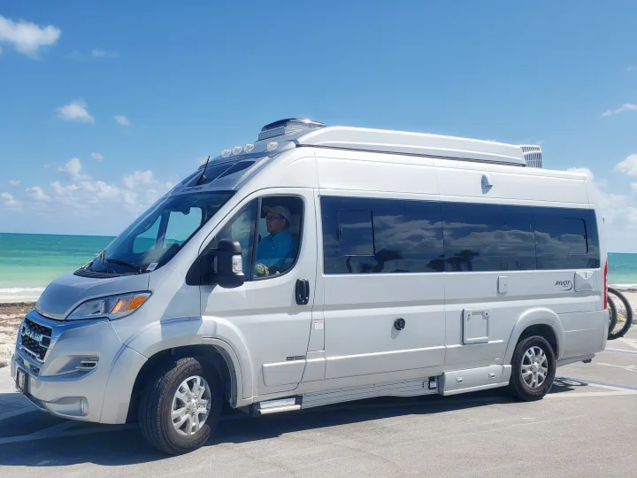 Silver RV rental parked oceanside, with a person in the driver's seat. Blue sky, turquoise water, and sandy beach are visible.. Roadtrek PIVOT SLUMBER 2024