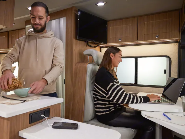 Inside an RV rental: a man pours cereal while a woman works on her laptop at a dinette.. Roadtrek PIVOT SLUMBER 2024