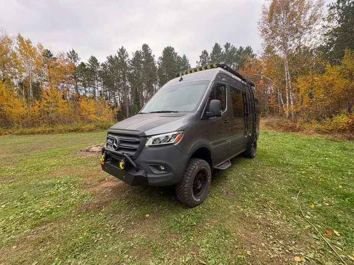 This gray Mercedes Sprinter adventure van, ideal for rv rental, is parked in a beautiful autumn forest clearing with fall foliage.. Storyteller Overland Storyteller Overland Class B 2024