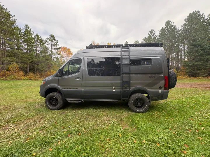 This gray Storyteller Overland MODE **rv rental**, an off-road camper van, is parked in a grassy forest clearing.. Storyteller Overland Storyteller Overland Class B 2024