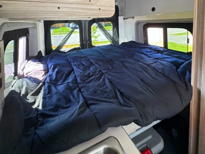 Cozy RV rental bed with a dark blue comforter, screens on windows, and a view of the street outside.. Winnebago Solis 59PX 2025