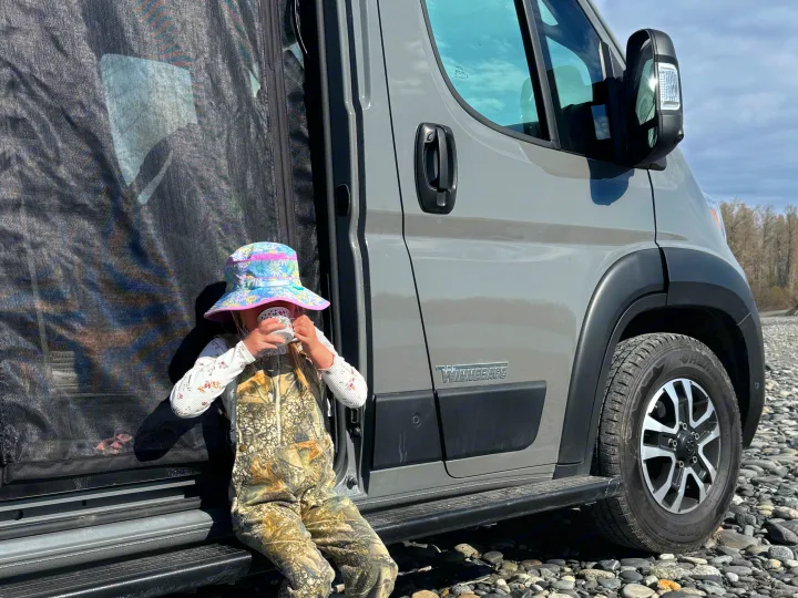 Little one sips from a mug next to a Winnebago RV rental van, parked on a rocky riverbed. Great for family adventures!. Winnebago Solis 59PX 2025