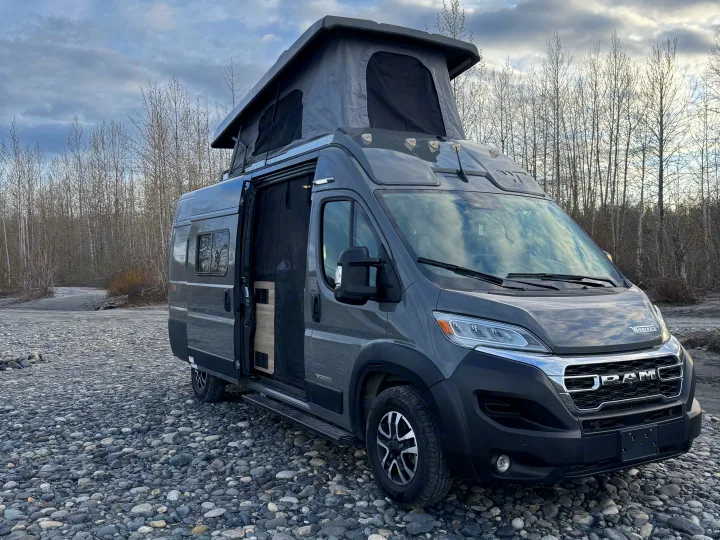 This gray pop-top camper van, possibly an RV rental, is parked on a rocky riverbed with bare trees in the background.. Winnebago Solis 59PX 2025