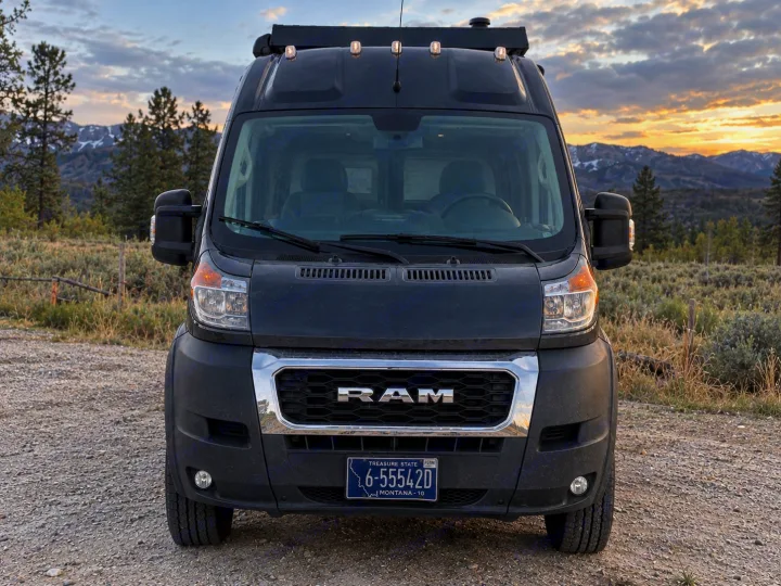 Black Ram rv rental camper van parked on gravel, front view. Mountains & trees under a cloudy sky, ideal for outdoor adventures.. Dodge Ram 1500 2020