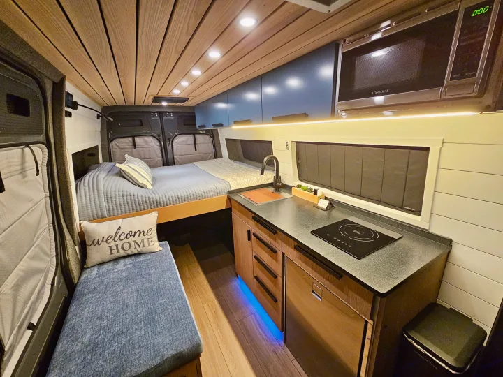 This modern rv rental interior features a cozy bed, full kitchenette, and sleek wooden accents, ideal for travel.. Mercedes-Benz Sprinter 2023