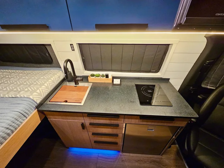 RV rental interior: A modern kitchenette with a sink, induction cooktop, cozy bed, and illuminated cabinetry.. Mercedes-Benz Sprinter 2023