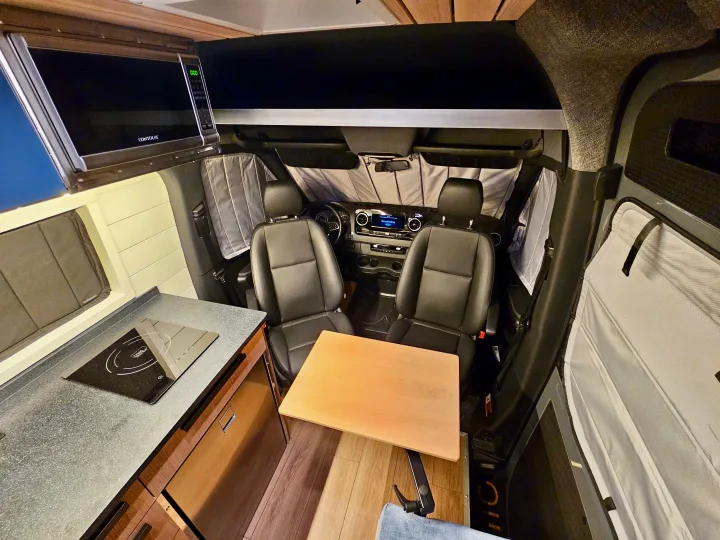 Inside this modern rv rental camper van: compact kitchen with microwave, induction hob, dining area, and front cabin seats.. Mercedes-Benz Sprinter 2023
