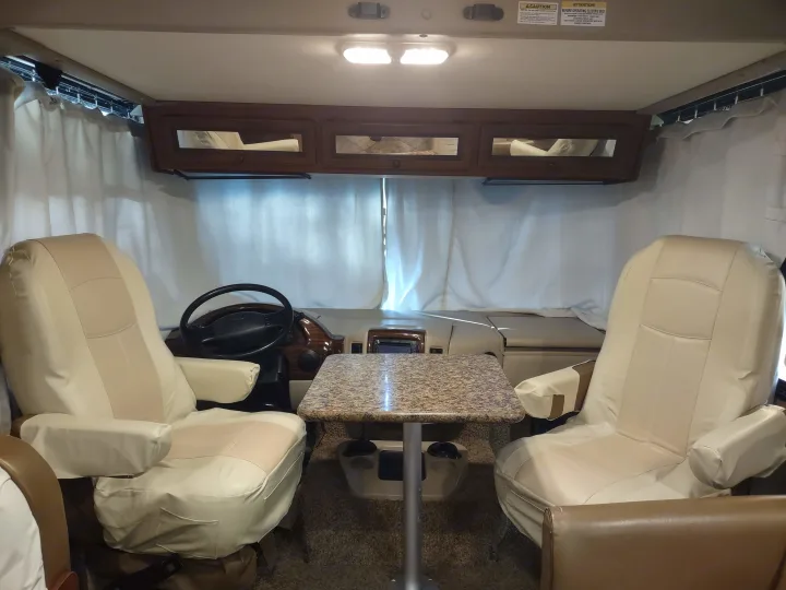 Interior of an RV rental showing protected beige captain's chairs, steering wheel, and a small table in the front cabin area.. Thor Windsport 2015