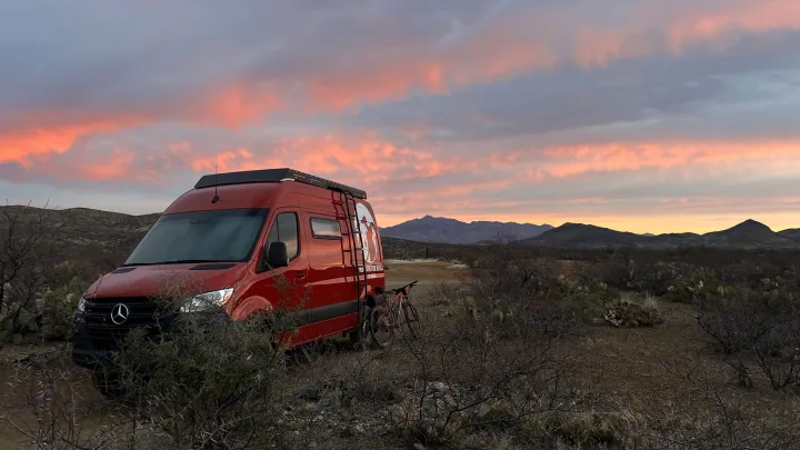 Red Mercedes-Benz Sprinter RV rental amidst a thorny desert landscape with colorful sunset clouds above mountains.. Mercedes-Benz Other 2022