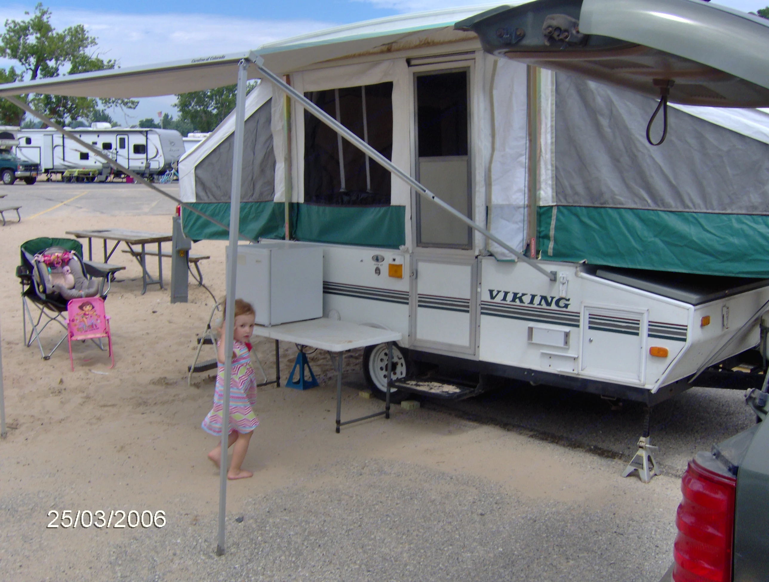 Family enjoying an RV rental. A child plays by a pop-up camper with awning, fridge, and table in a campground.. Viking 1906 2001