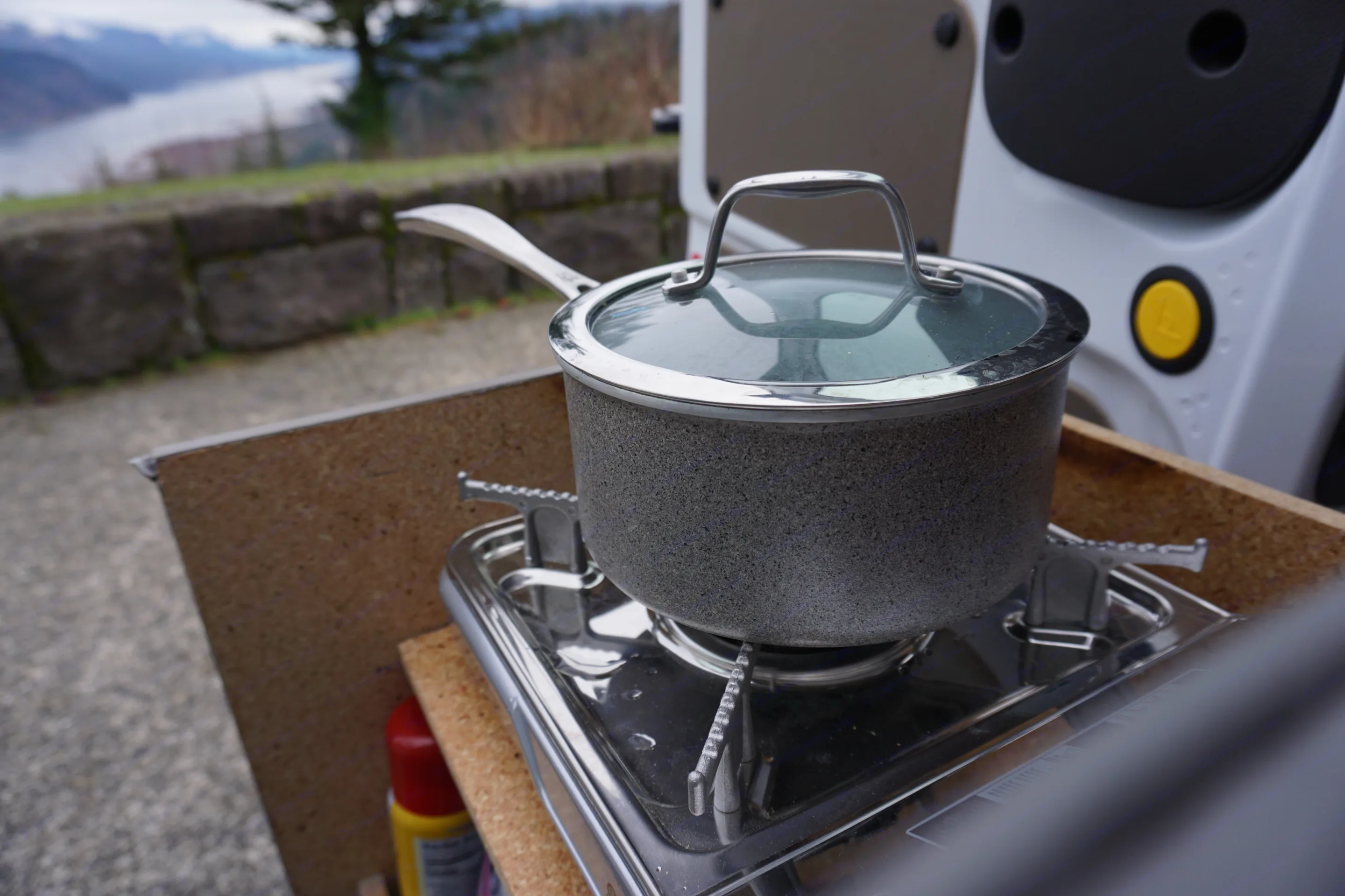 A speckled gray pot with a glass lid sits on a portable gas stove, part of an RV rental kitchen setup with a scenic outdoor view.. Ford Transit Custom 2017