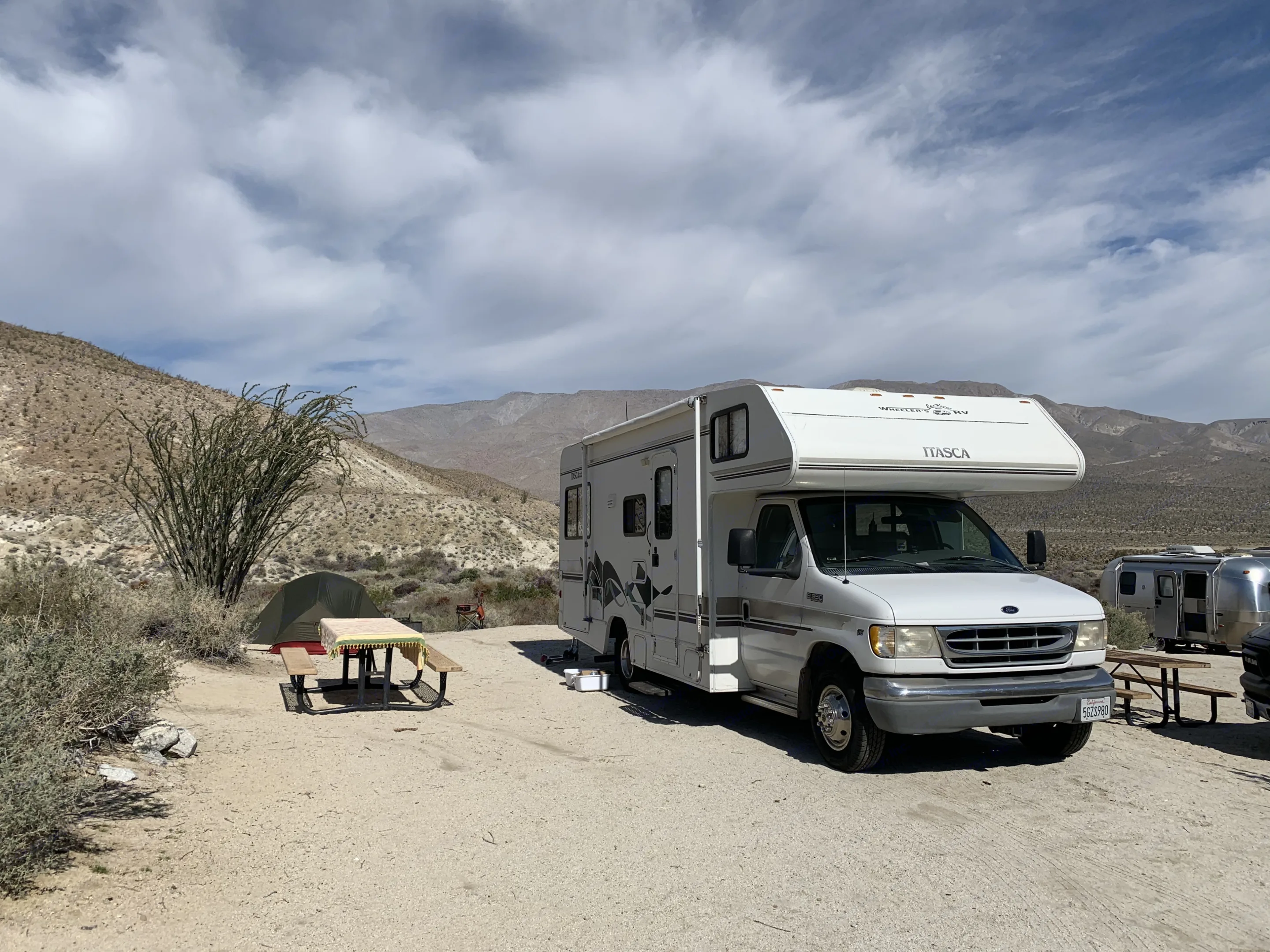 A white Itasca rv rental rests in a dry desert campsite with a tent, picnic table, and Ocotillo plant. Mountains loom under clouds.. Itasca Spirit 1999