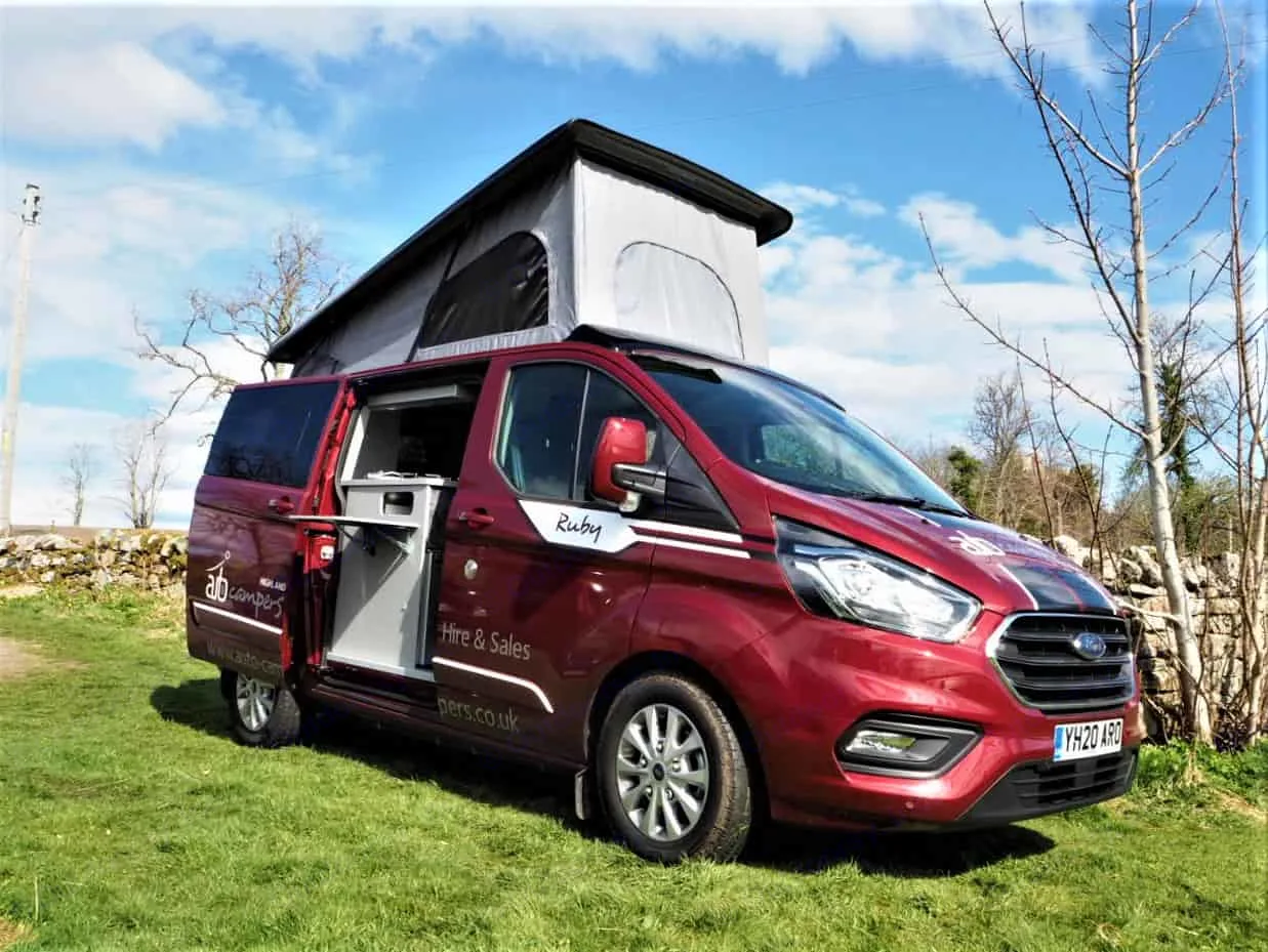 This red Ford Transit Custom campervan with pop-top roof is parked on grass. Its side door is open, perfect for RV rental adventures.. Ford Transit Custom 2+2 berth Pop Top, manual 2020