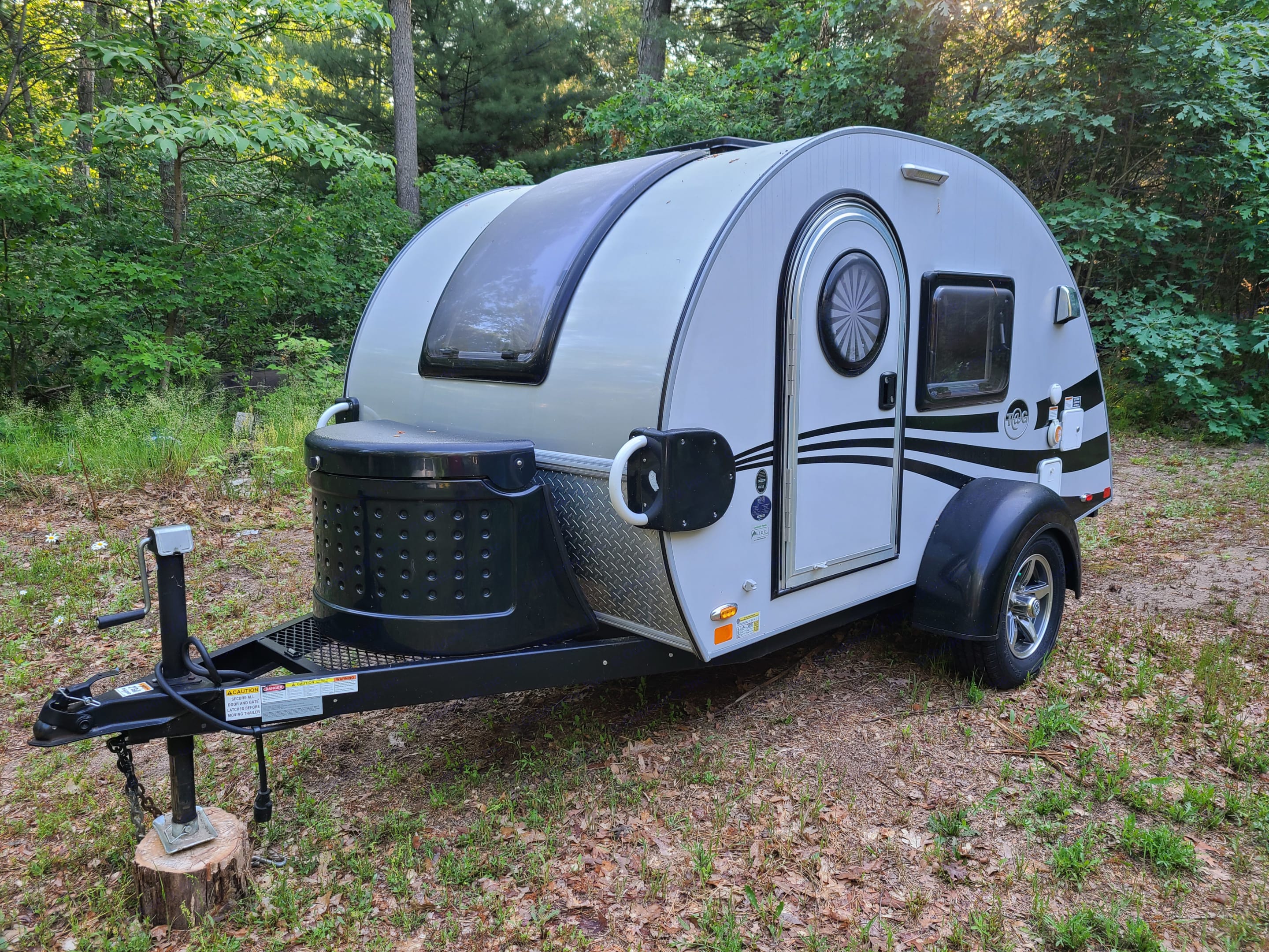 This compact RV rental is a white teardrop trailer parked in a serene wooded campsite.. Nucamp Tag 2018
