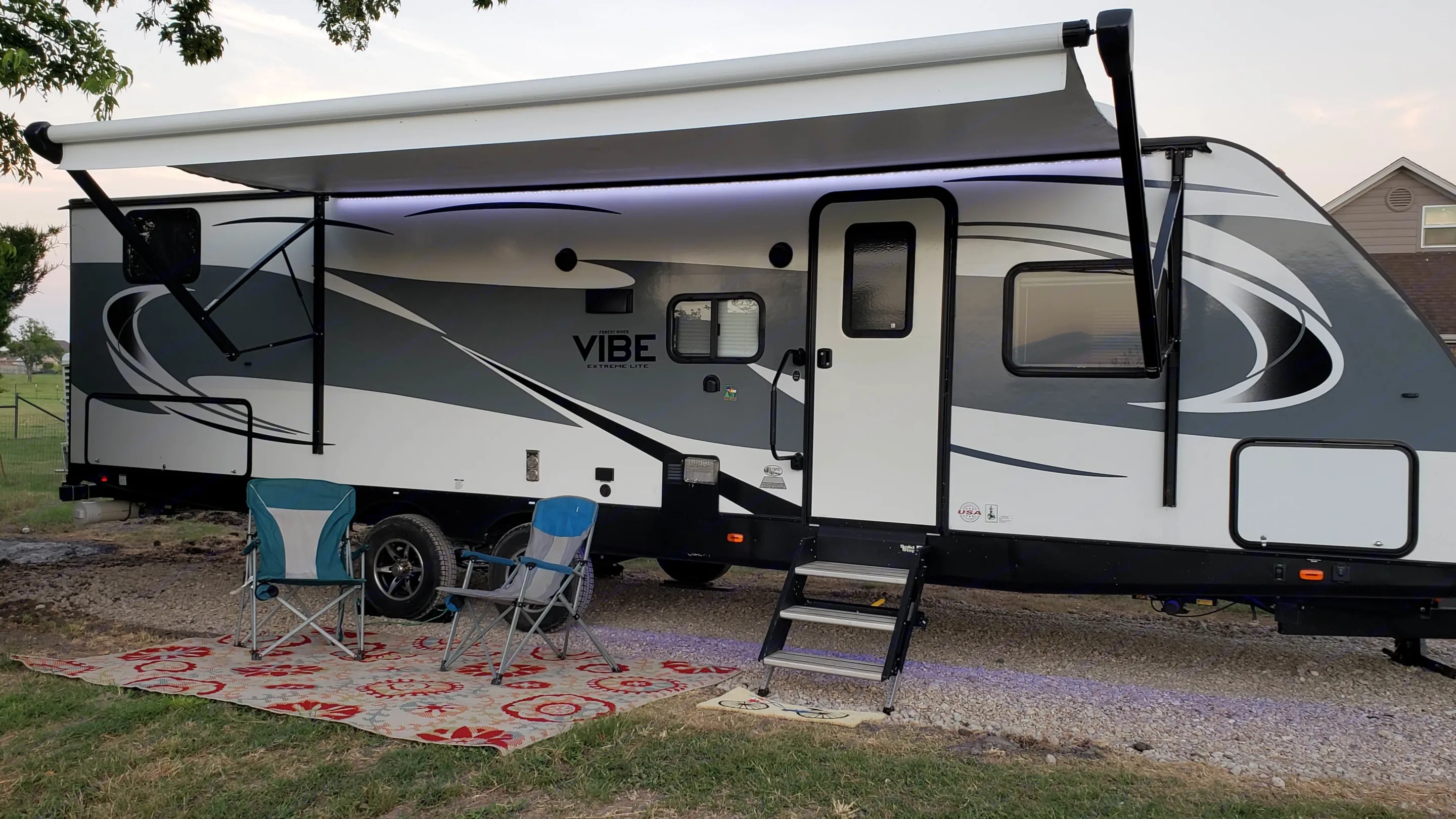 Relaxing RV rental setup: a white and gray Vibe travel trailer with an extended awning, two chairs, and a mat outside.. Forest River Vibe 2019