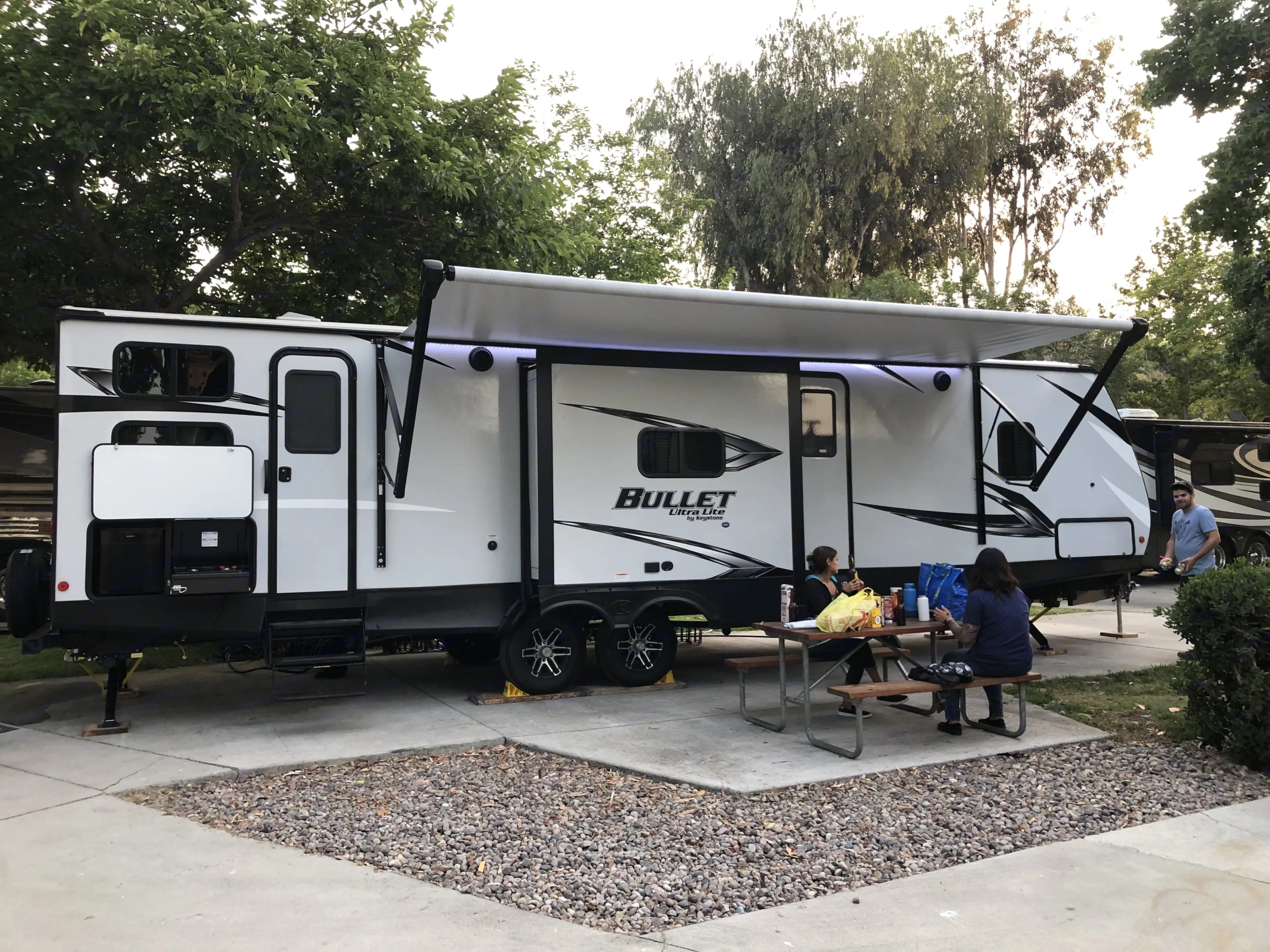 RV rental: A white 'Bullet' travel trailer with extended awning. People enjoy a picnic at their campground site.. Keystone Bullet 2021