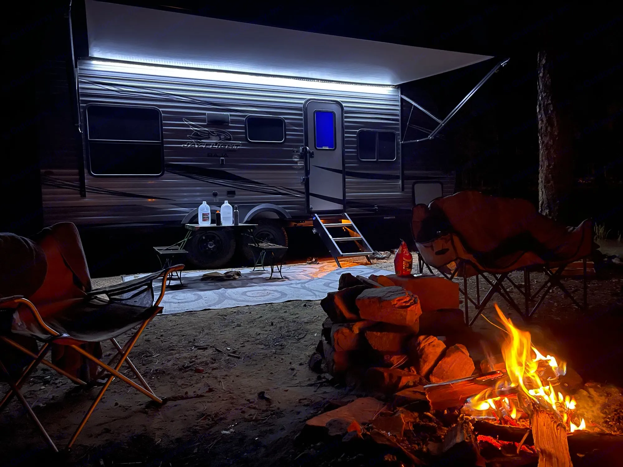 Serene night camping scene with an RV rental, illuminated awning, and a crackling campfire surrounded by chairs.. Jayco Baja 2021