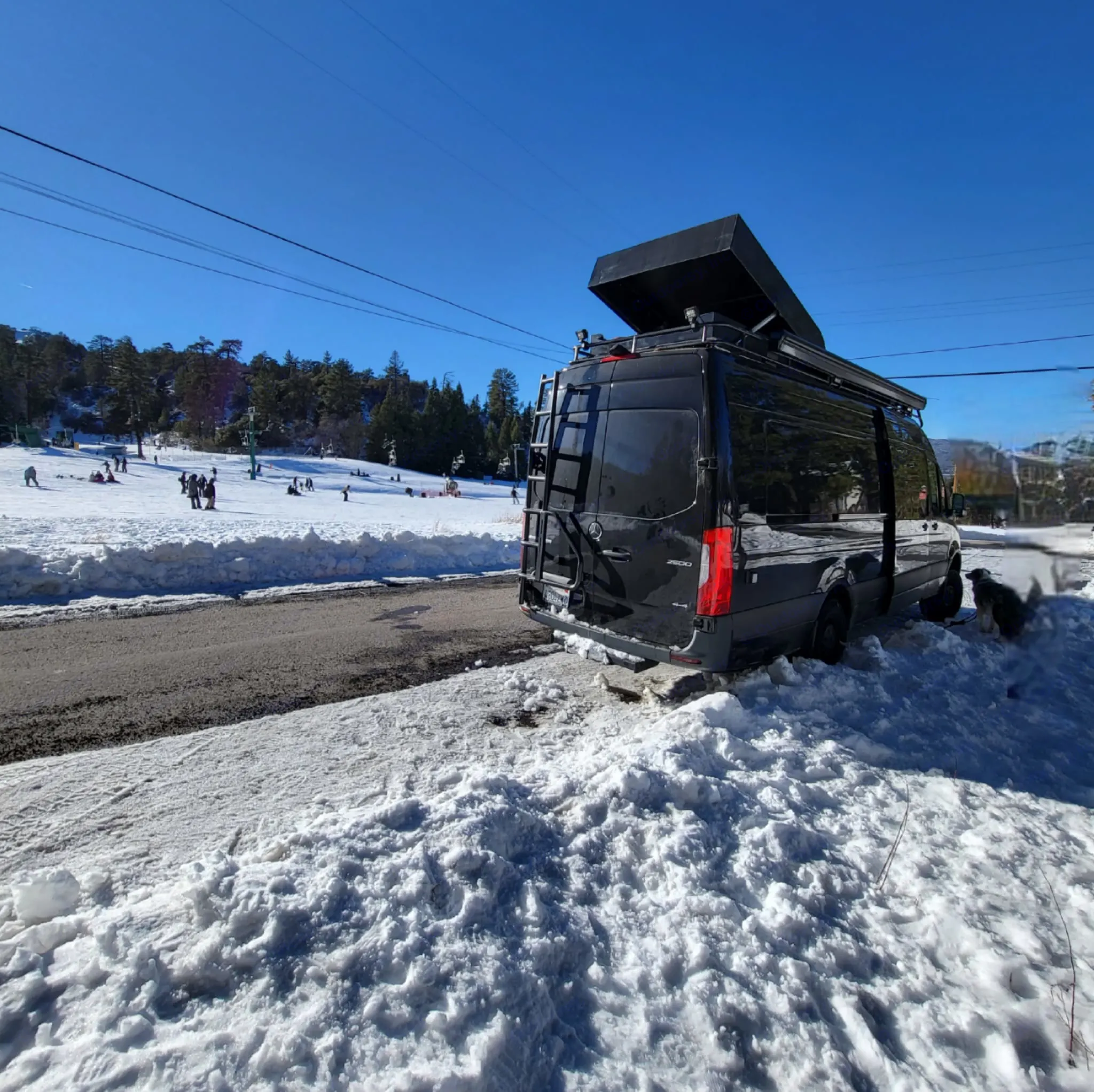 Black camper van (RV rental) parked in snow by a ski slope with people enjoying winter activities. A dog is leashed.. Mercedes-Benz Sprinter 2019