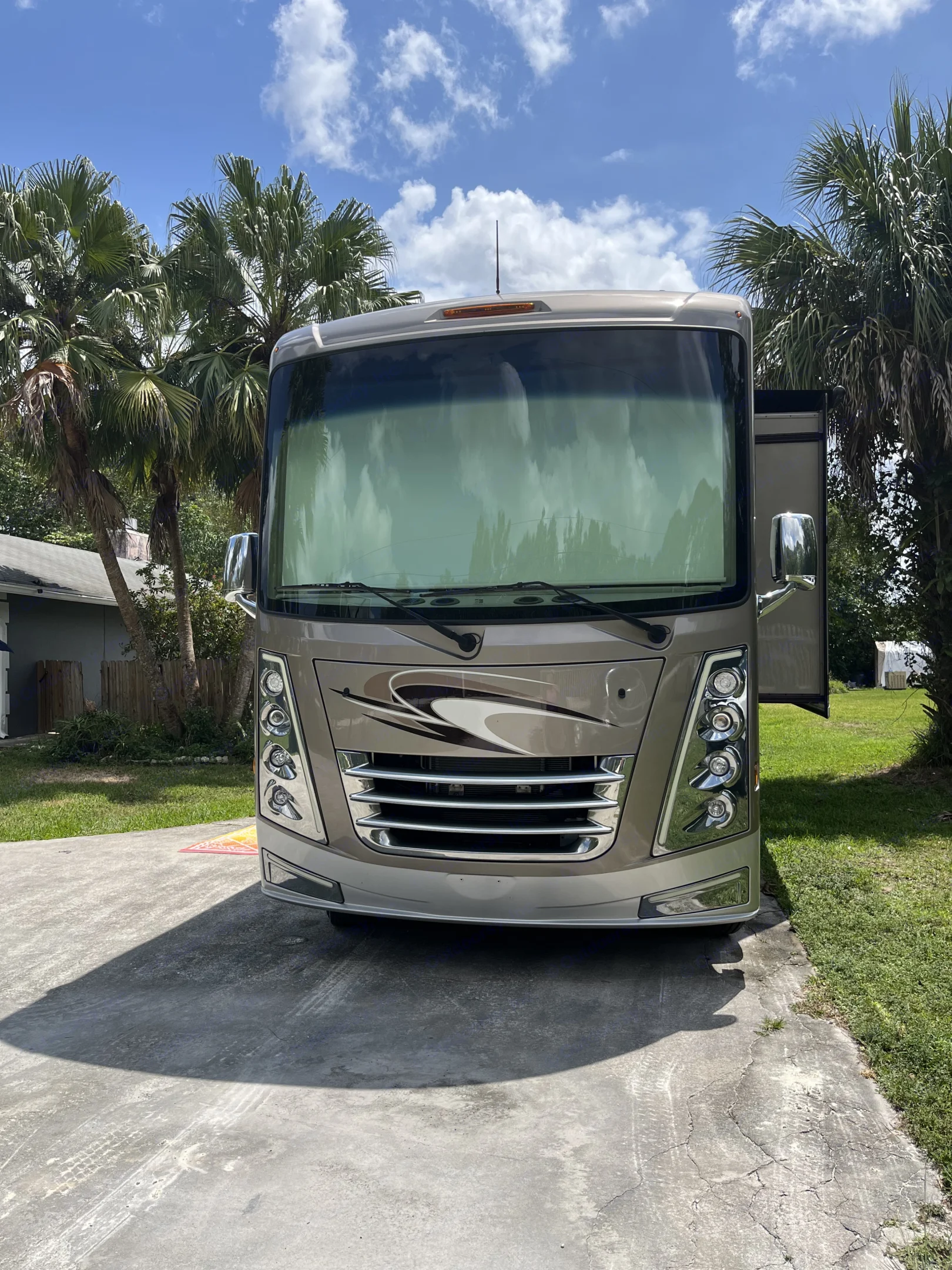 Front view of a sleek tan RV on a sunny driveway, surrounded by palm trees.. Thor Challenger 2021