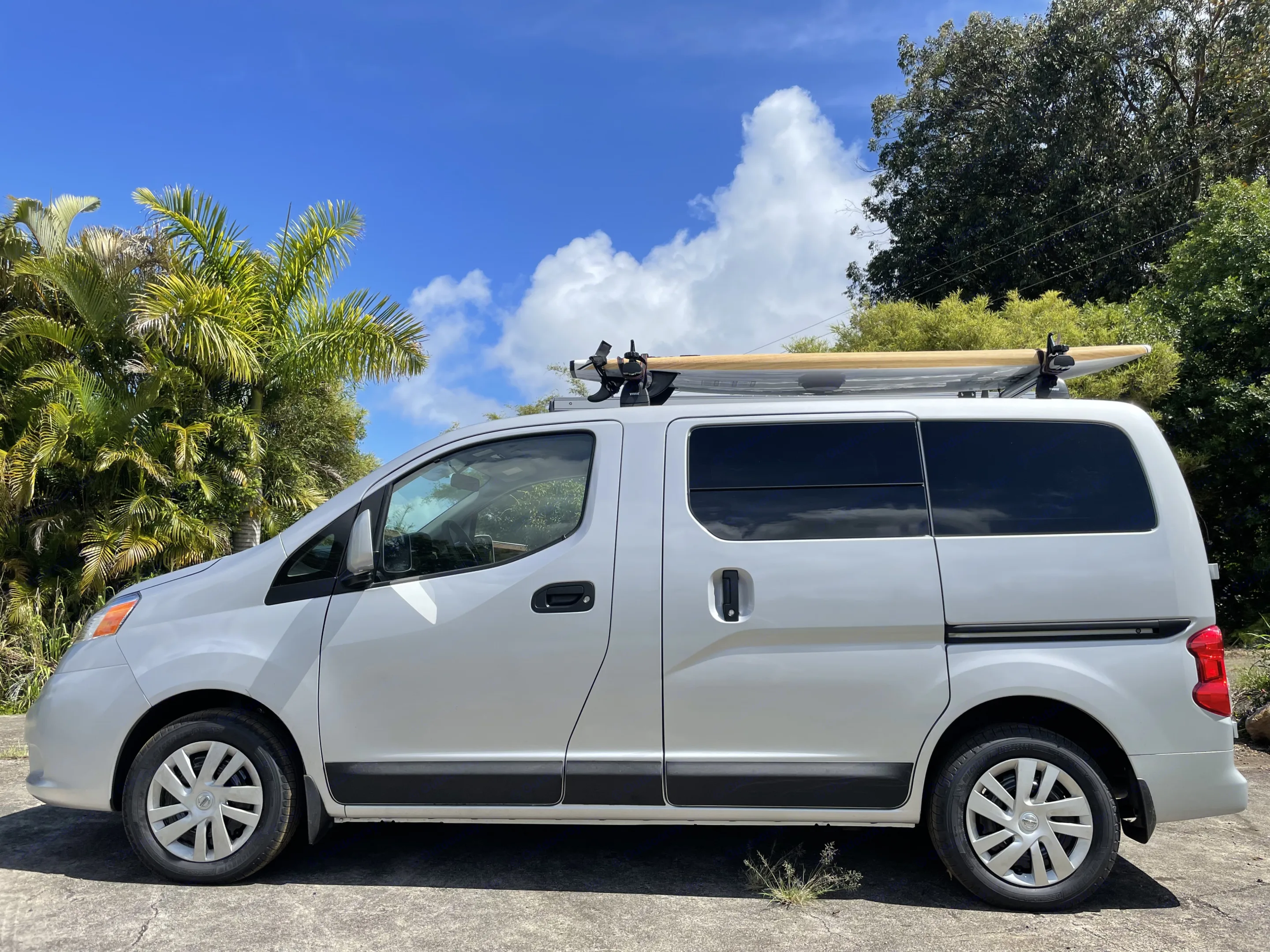 Compact RV rental: A white van with a surfboard on its roof, parked under a blue sky with vibrant palm trees, ready for adventure.. Nissan NV200 2021