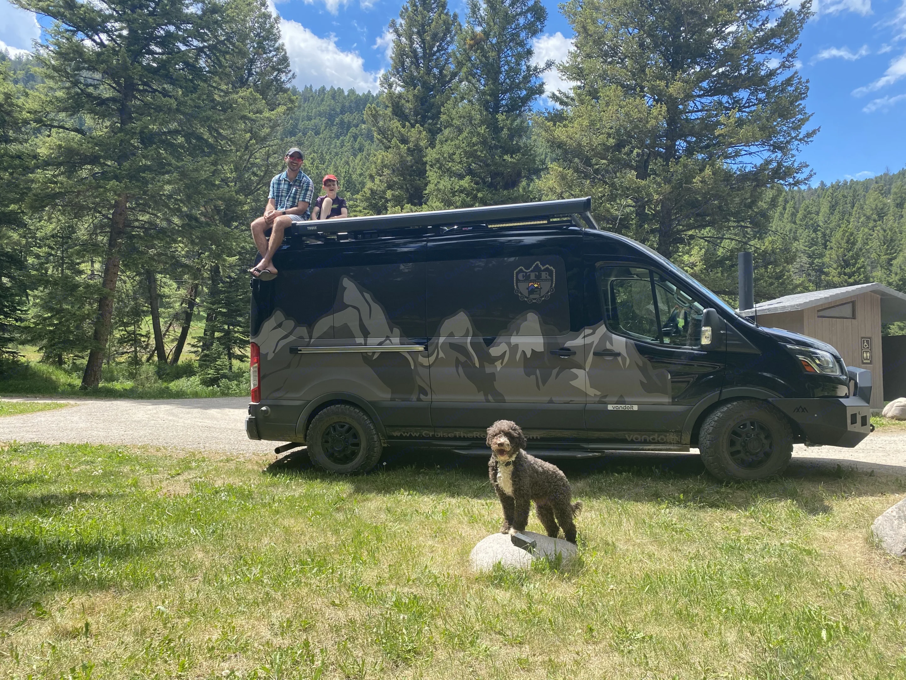 Family enjoying their black RV rental. Dad & son on the roof, dog on a rock, surrounded by green forest & mountains.. Ford Custom Transit 2020