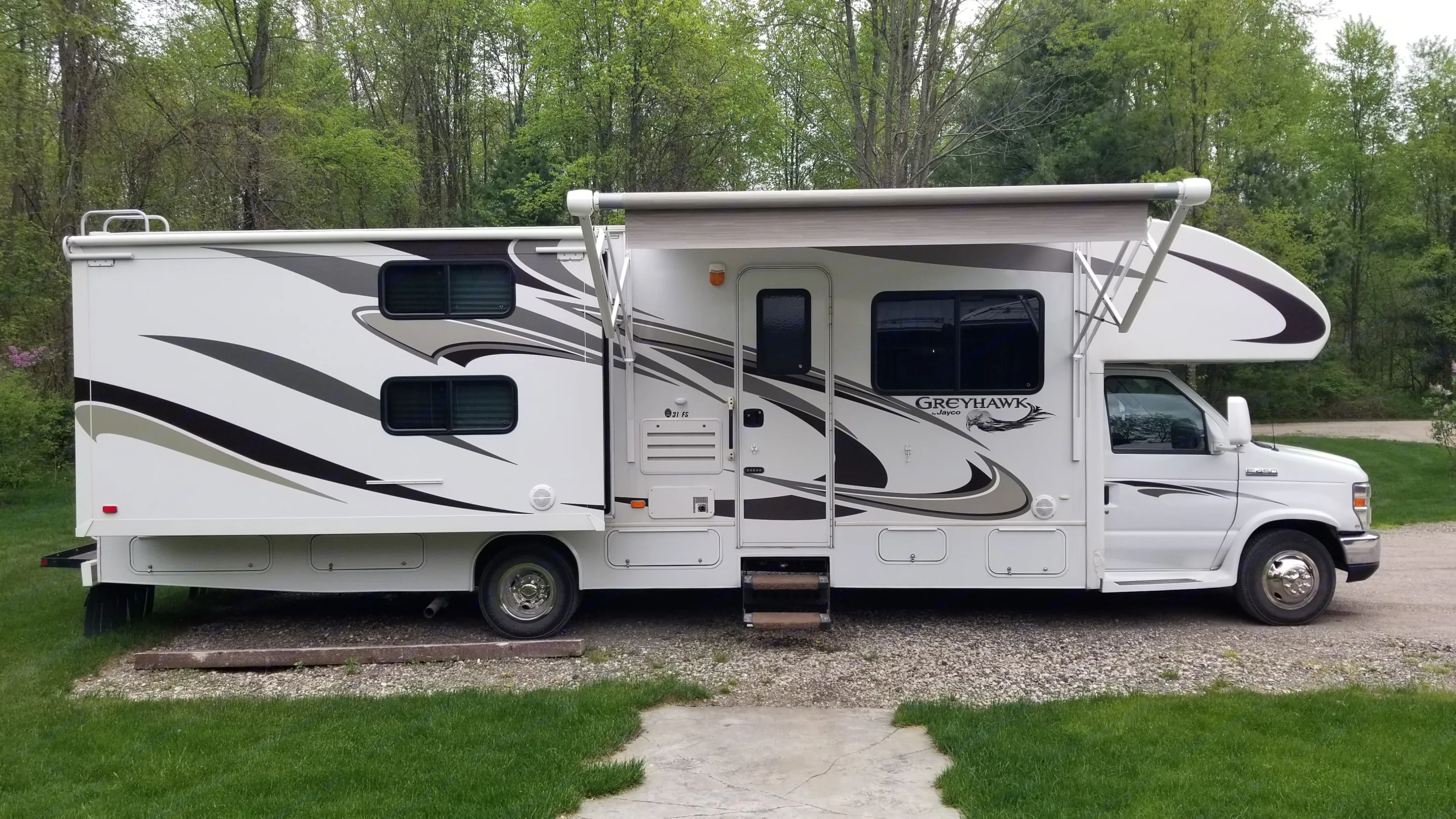 White Jayco Greyhawk RV with extended awning, parked on gravel near trees. A great option for your next rv rental adventure.. Jayco Greyhawk 2013