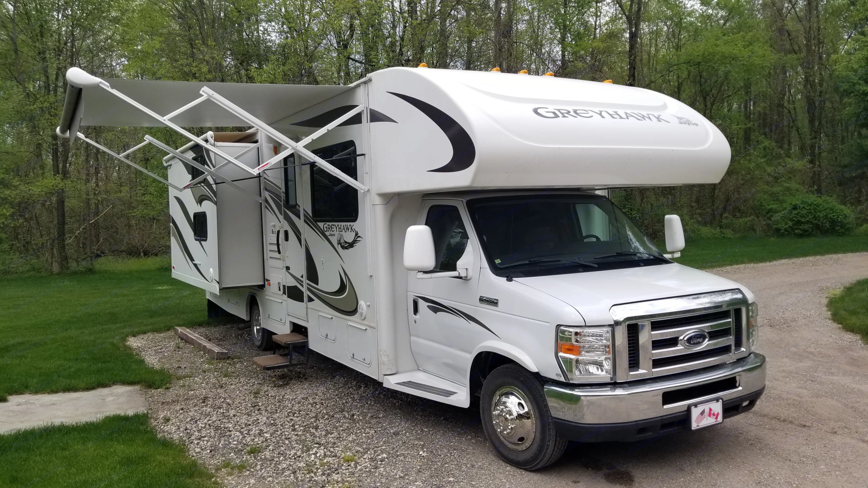 White Class C **RV rental** with awning extended and slide-out open, parked on gravel beside green grass and trees.. Jayco Greyhawk 2013